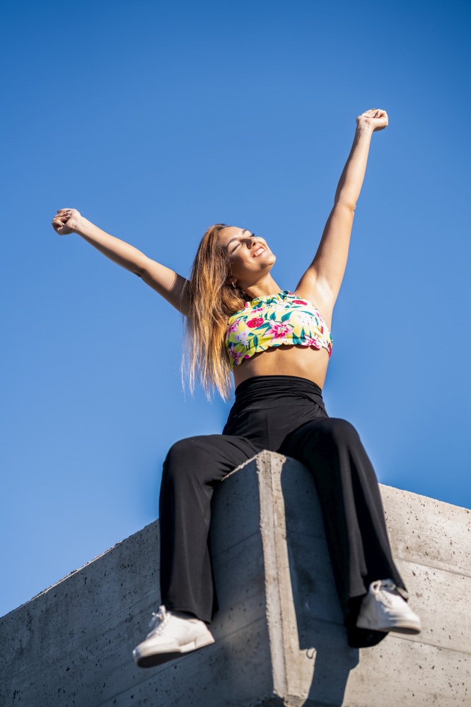 Young woman opening arms against blue sky sitting on urban wall.. Young woman opening arms against blue sky