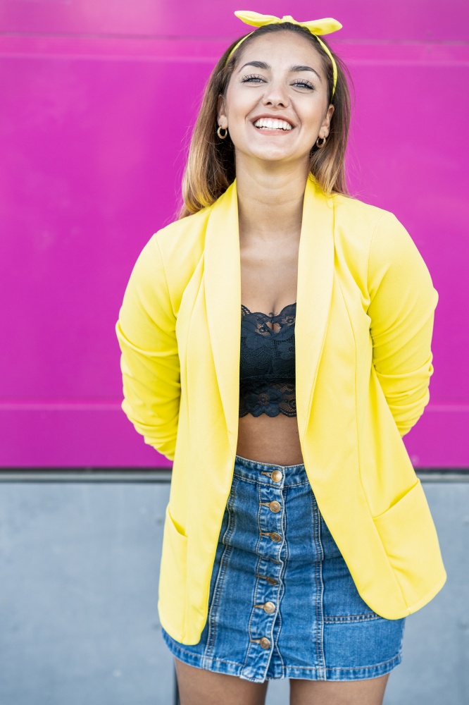 Happy woman wearing a yellow jacket and headband, near an urban purple wall. Young woman wearing a yellow jacket and headband