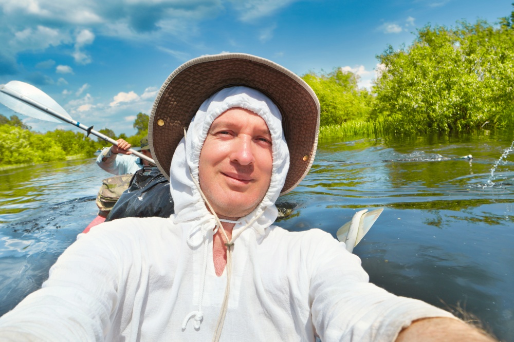 Smiling man in white hoodie and hat taking selfie photo on kayak during summer river adventure trip