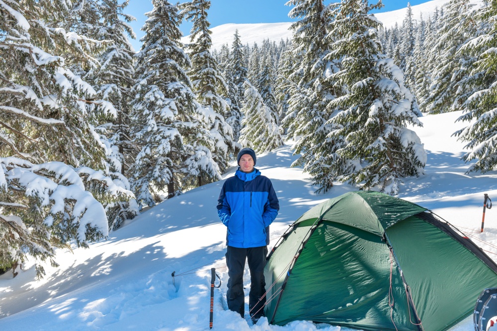 Man in forest in winter mountains with deep snow standing near tent and looking into camera
