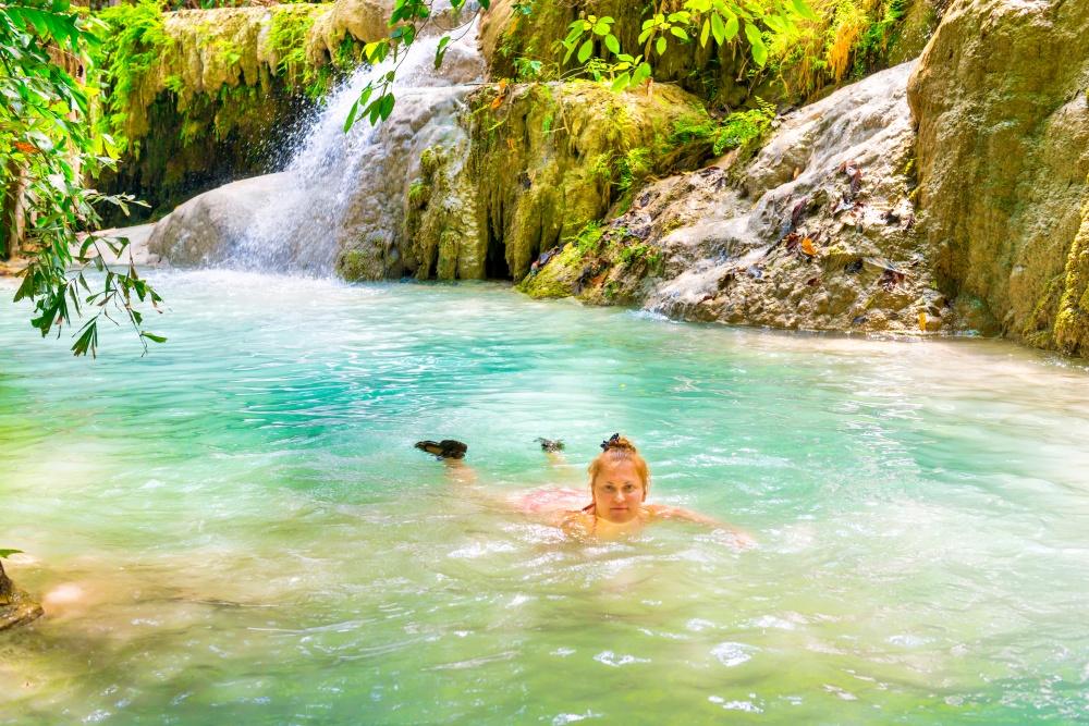Red haired young woman in pink bikini swimsuit swims in emerald blue water of tropical lake with waterfall. Erawan National park, Kanchanaburi, Thailand