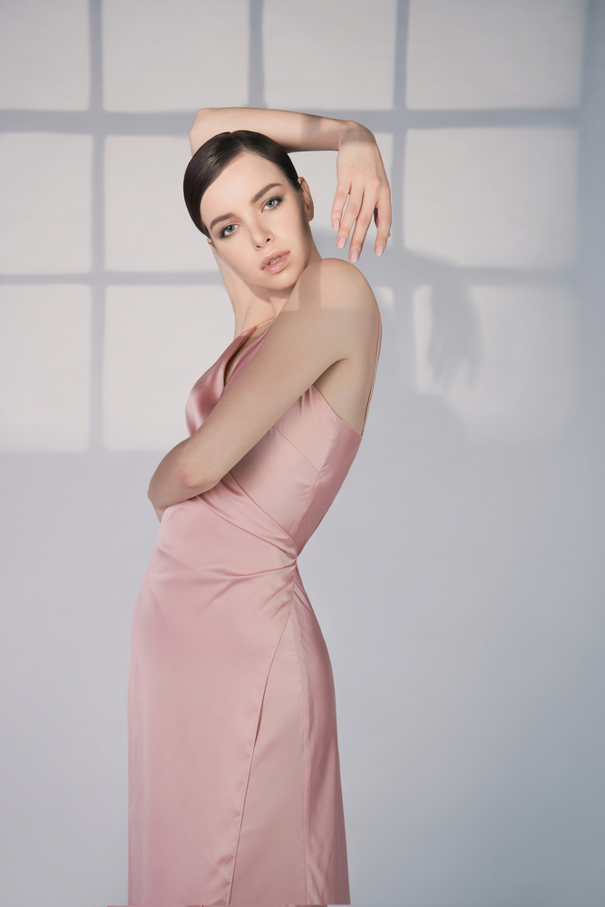Woman in elegant fashionable dress. Beautiful model pose in studio in pink evening clothes. Classic silk dress. Sexy lady stay on white background. Perfect young woman with classic hairstyle and makeup.