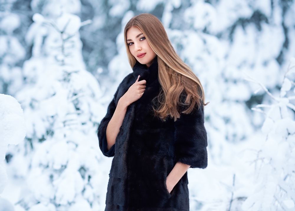 Fashion young woman in the winter forest. Young elegant model in black fur coat in nature. Blonde girl with long smooth hair