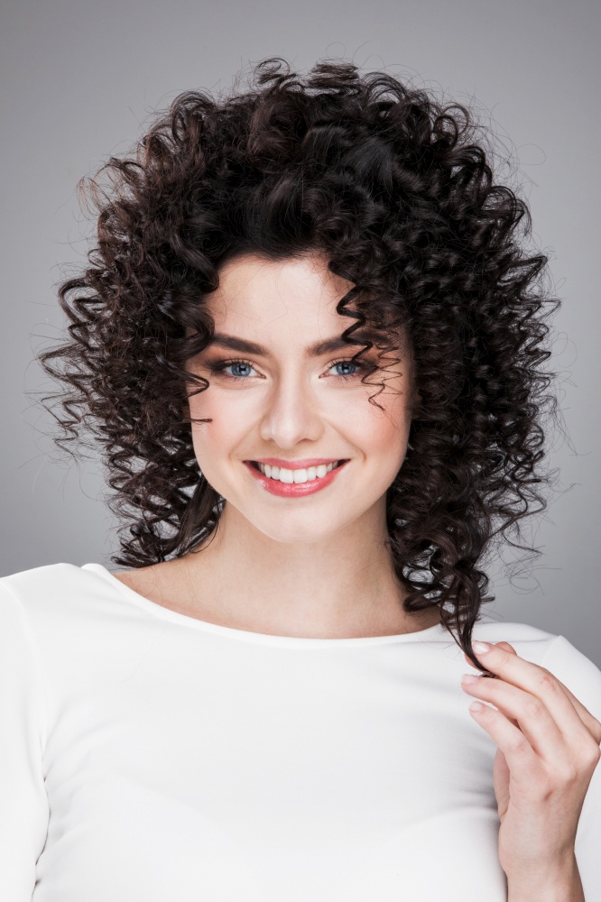 Beauty young woman portrait on gray background. Girl touching her black curly hair. Fresh Clean Skin and Blue Eyes. Hairstyle. Haircut. Model girl touching her curly hair