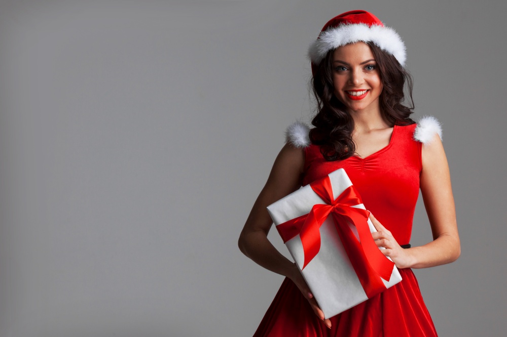 Smiling cute girl in red christmas outfit holding gift box on gray background. Girl with christmas gift box