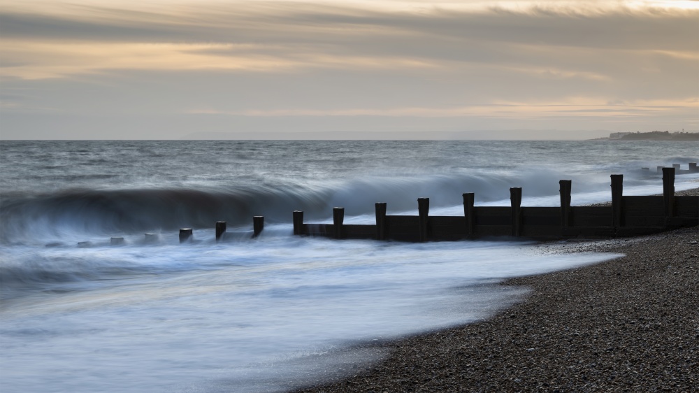 Beautiful abstract long exposure landscape image of waves crashing onto groynes on beach during sunset