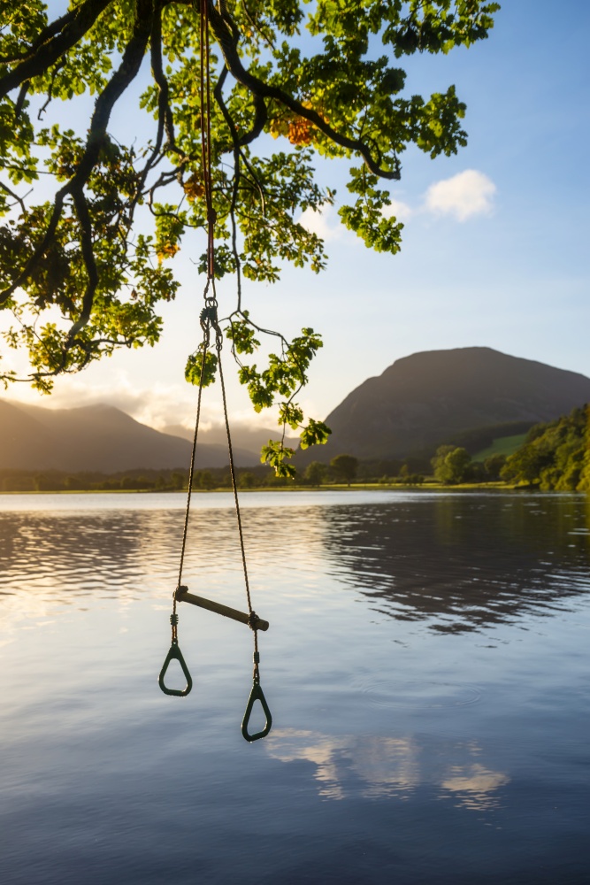 Quintessential English Summer landscape image of child&rsquo;s rope swing over calm lake in Lake District during golden Summer sunrise