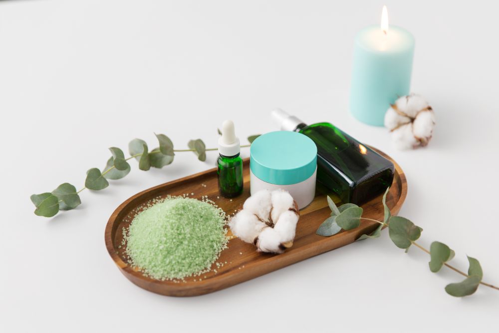 beauty and spa concept - green bath salt, serum with dropper, body oil, moisturizer and eucalyptus cinerea with cotton flowers on wooden tray. bath salt, serum, moisturizer and oil on tray