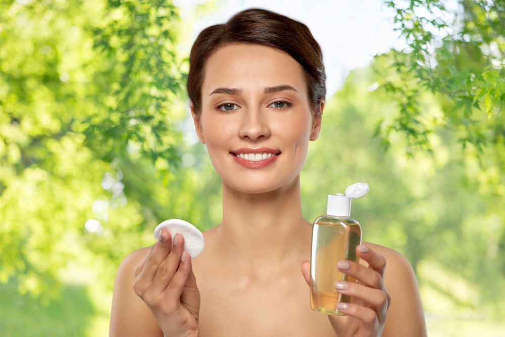 beauty, skin care and people concept - smiling young woman with toner or cleanser and cotton pad over green natural background. young woman with toner or cleanser and cotton pad