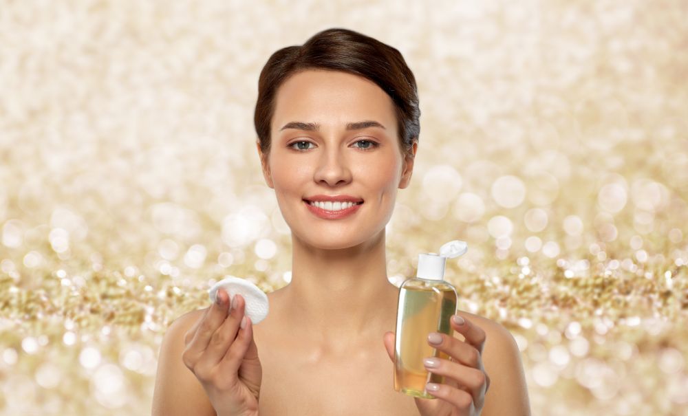 beauty, skin care and people concept - smiling young woman with toner or cleanser and cotton pad over shimmering golden glitter on background. young woman with toner or cleanser and cotton pad