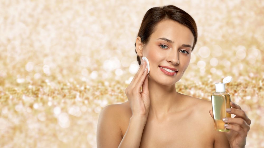 beauty, skin care and people concept - smiling young woman with toner or cleanser and cotton pad cleansing face over shimmering golden glitter on background. young woman with toner or cleanser and cotton pad