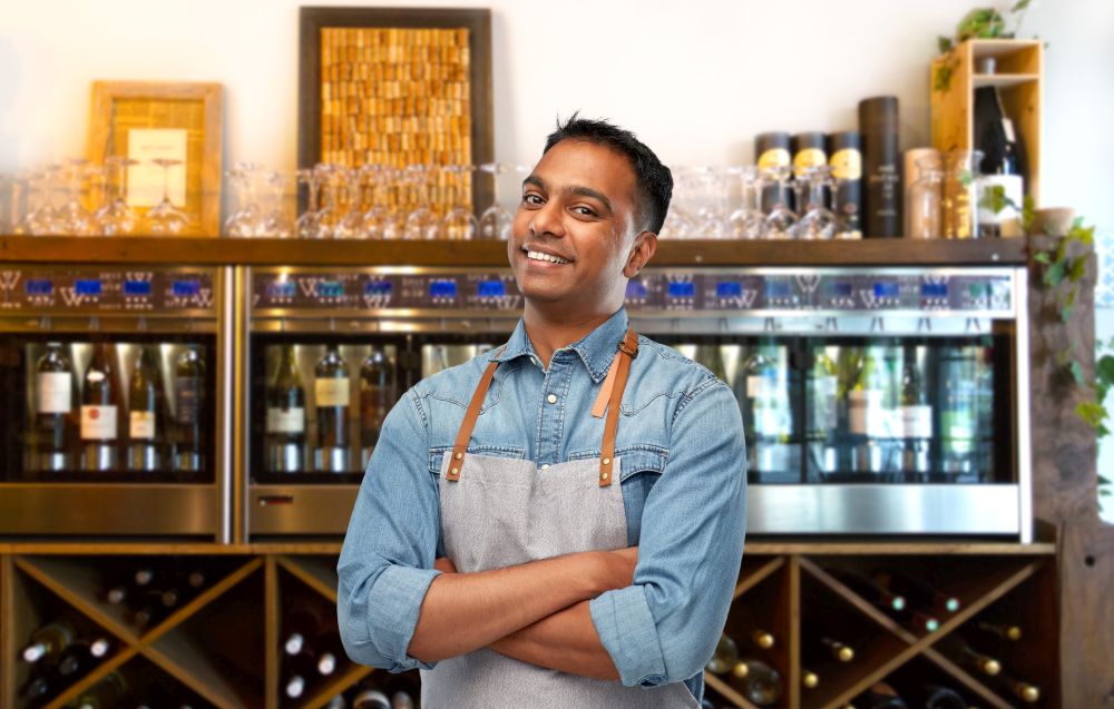people, job and profession concept - smiling indian barman, waiter or salesman in apron over wine bar background. smiling indian barman or waiter in apron at bar