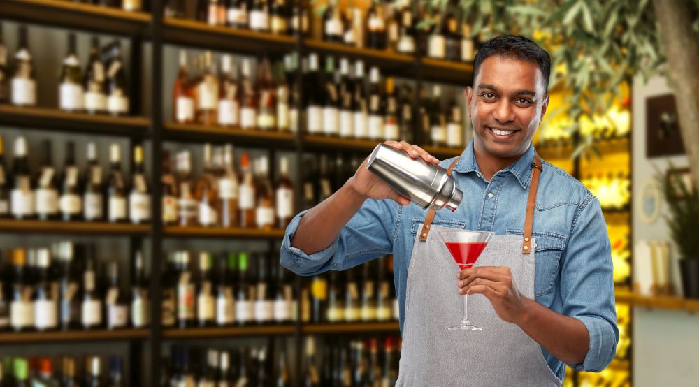 alcohol drinks, people and profession concept - indian barman in apron pouring cocktail from shaker to glass over bar background. indian barman with glass of cocktail and shaker
