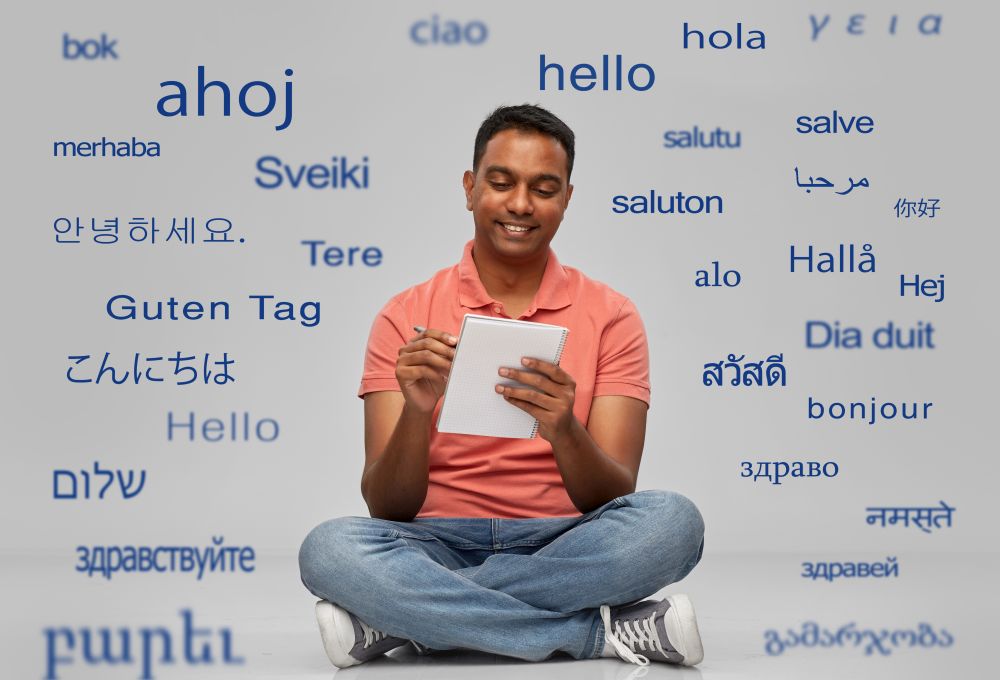 communication, learning and people concept - smiling indian man in polo shirt writing to notebook sitting on floor over greeting words in different foreign languages on grey background. indian man in polo shirt writing to notebook