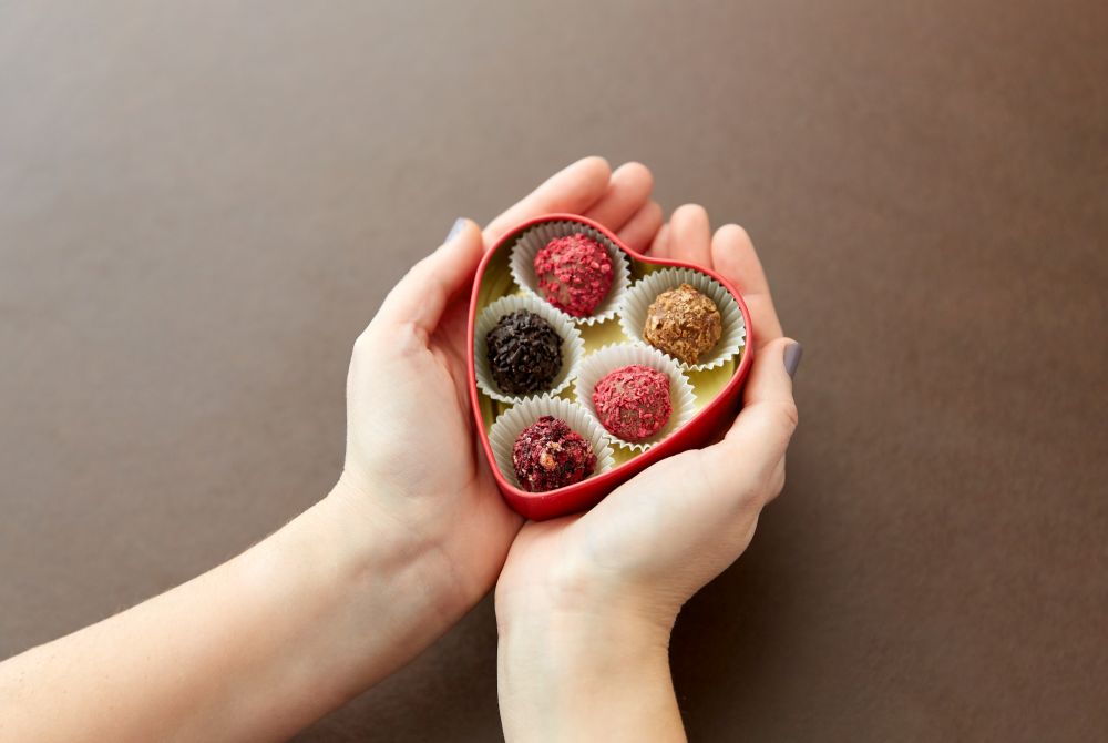 sweets, confectionery and food concept - hands holding candies in red heart shaped chocolate box on brown background. hands with candies in heart shaped chocolate box
