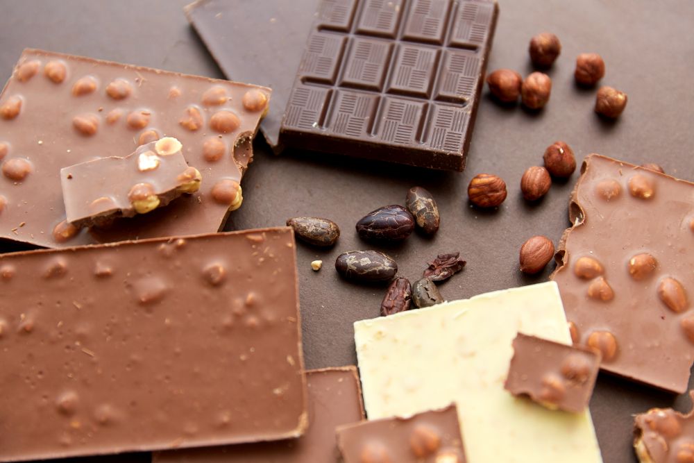 sweets, confectionery and food concept - milk, dark and white chocolate bars with hazelnuts and cocoa beans on brown background. chocolate bars with hazelnuts and cocoa beans