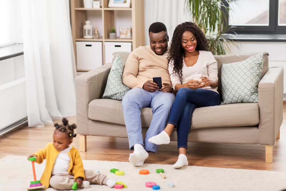 childhood and people concept - little african american baby girl playing with toy blocks and parents using smartphones at home. african baby girl playing with toy blocks at home