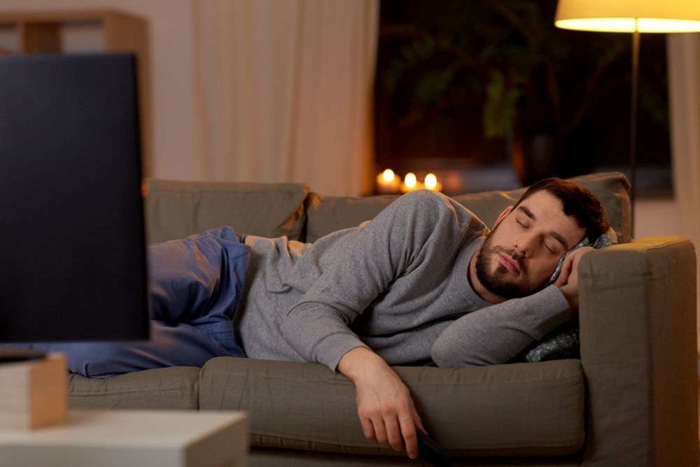 leisure and people concept - man sleeping on sofa with tv on at home at night. man sleeping on sofa with tv remote at home