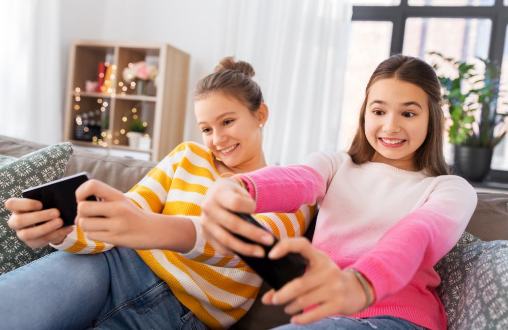 people, technology and friendship concept - happy teenage girls with smartphones sitting on sofa and playing game at home. girls and playing game on smartphones at home