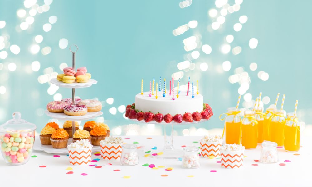 party and festive concept - birthday cake with candles and strawberries, drinks and food on table over lights on blue background. food and drinks on table at birthday party