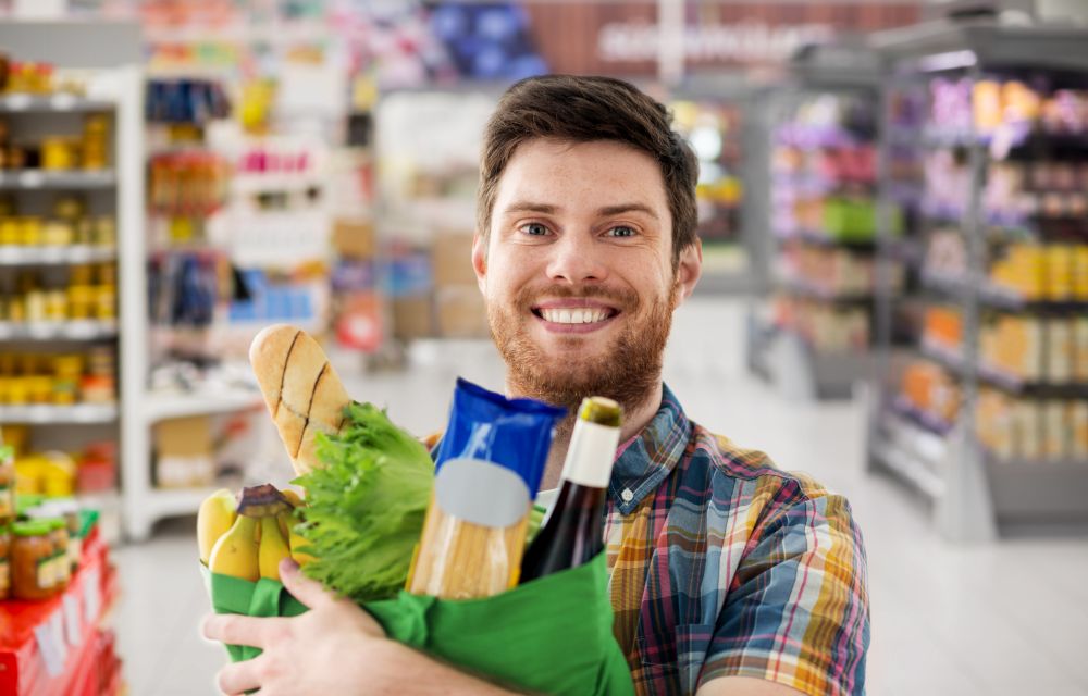 shopping, consumerism and people concept - smiling young man with food in bag over supermarket background. smiling young man with food in bag at supermarket