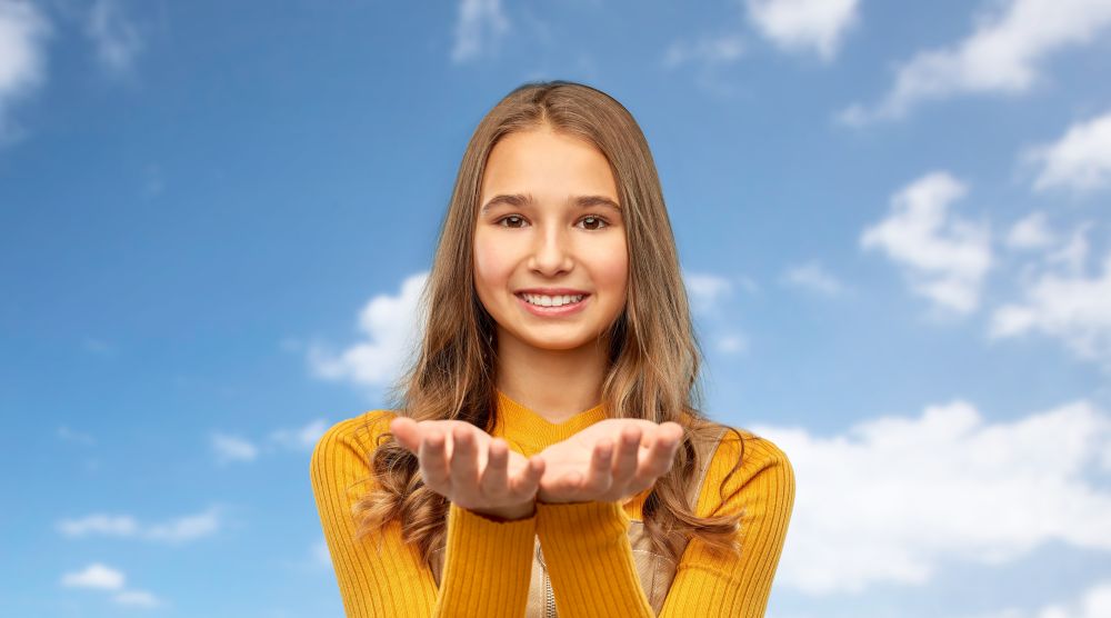 presentation and people concept - happy smiling young teenage girl holding something on empty hands over blue sky and clouds background. teenage girl holding something on empty hands