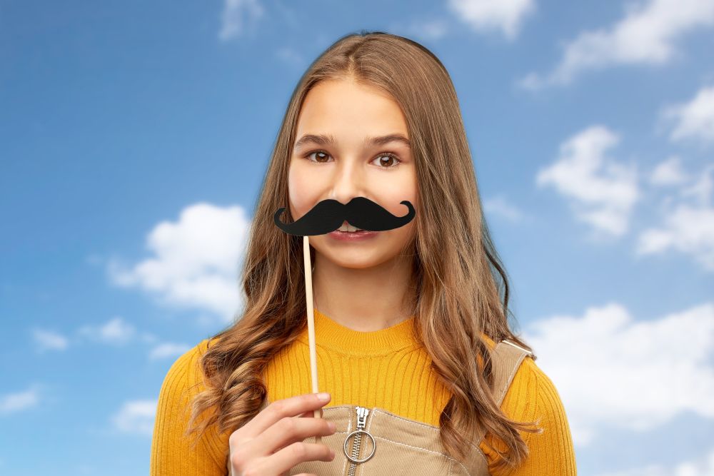 party props, photo booth and people concept - teenage girl with big black moustaches over blue sky and clouds background. teenage girl with black moustaches party accessory
