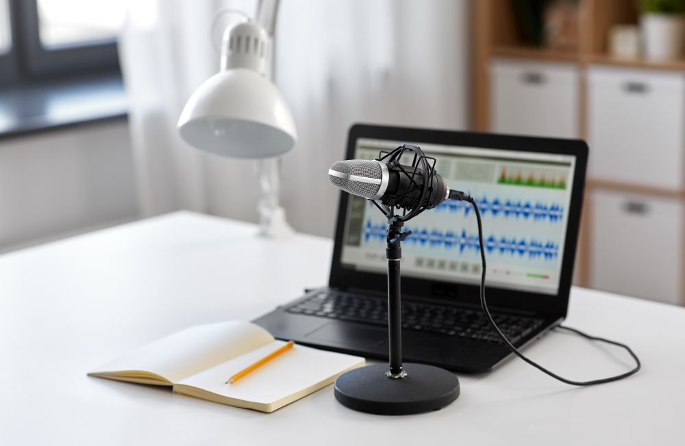post production and technology concept - microphone, laptop computer with sound editor program, headphones and notebook on table at home office. microphone, laptop and notebook on table