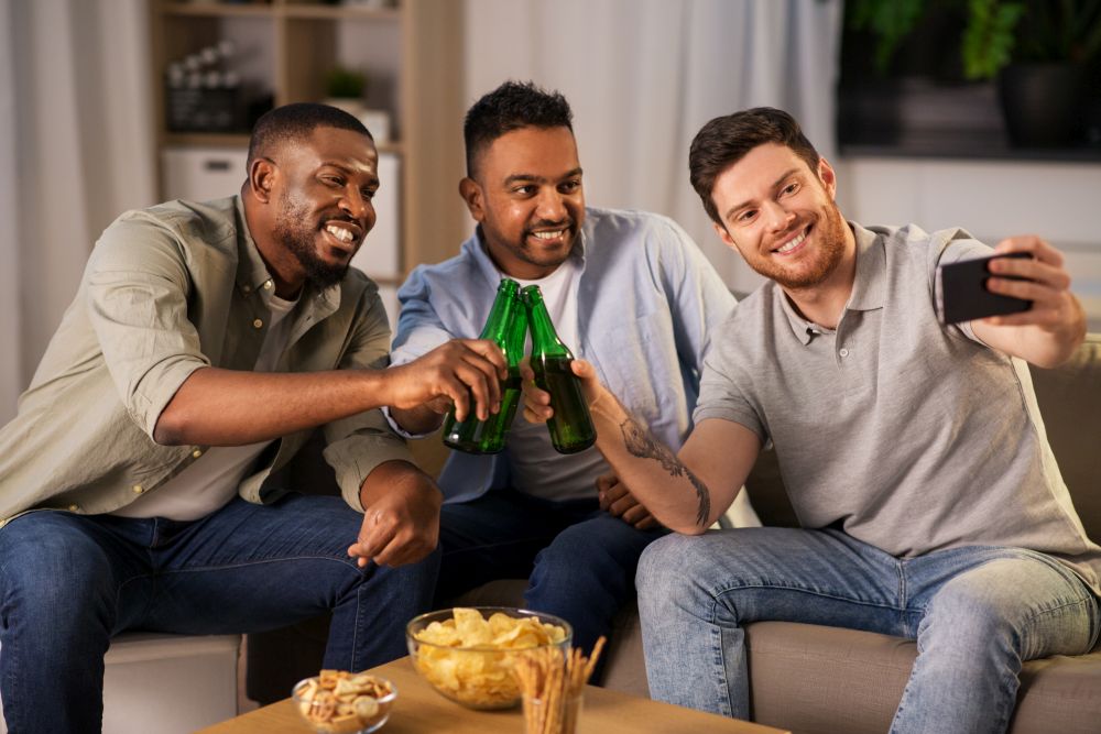 friendship, leisure and people concept - male friends with smartphone taking selfie and drinking beer at home at night. male friends with smartphone taking selfie at home