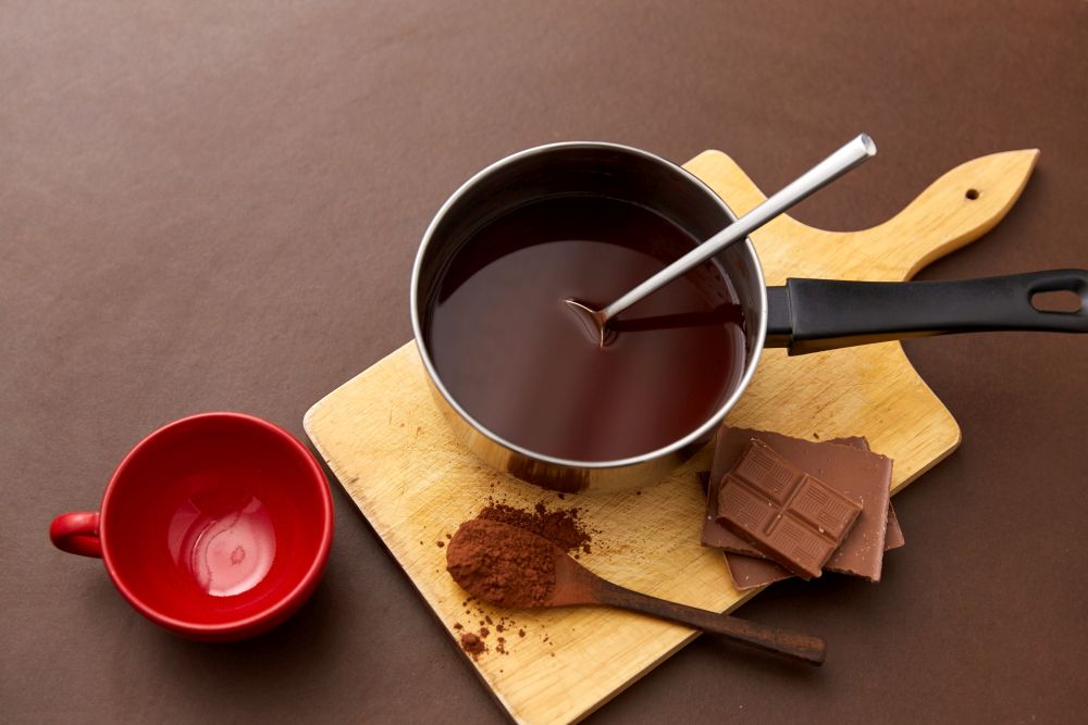 sweets, confectionery and culinary concept - pot with melted hot chocolate, cocoa powder in spoon, red ceramic mug and wooden board on brown background. pot with hot chocolate, mug and cocoa powder