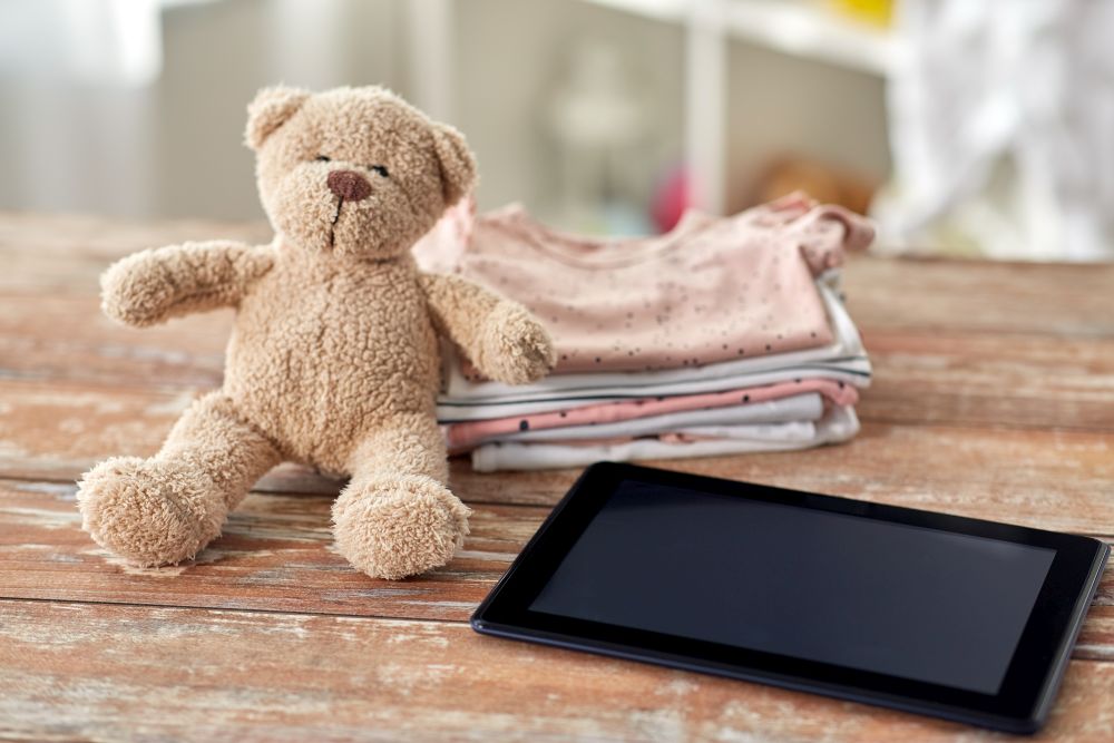 babyhood, technology and clothing concept - baby clothes, teddy bear toy and tablet pc computer on wooden table at home. baby clothes, teddy bear toy and tablet computer