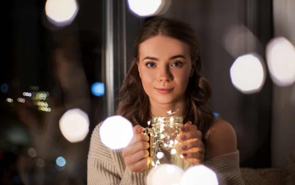 christmas, holidays and people concept - young woman in glasses with garland lights in mason jar mug at home at night. woman with christmas garland lights in glass mug