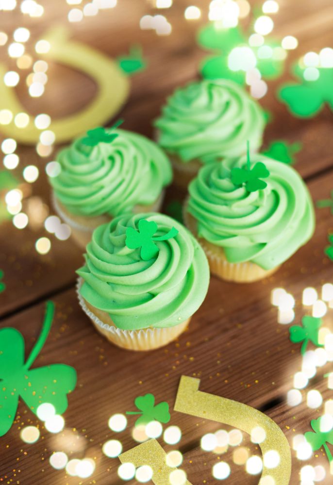 st patricks day, holidays and cooking concept - green cupcakes, horseshoes and shamrock on wooden table over festive lights. green cupcakes, horseshoes and shamrock