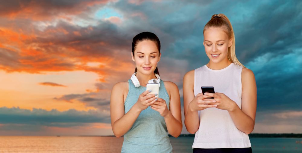 fitness, sport and healthy lifestyle concept - smiling young women or female friends with smartphones over sea and sunset sky on background. women or female friends with smartphones