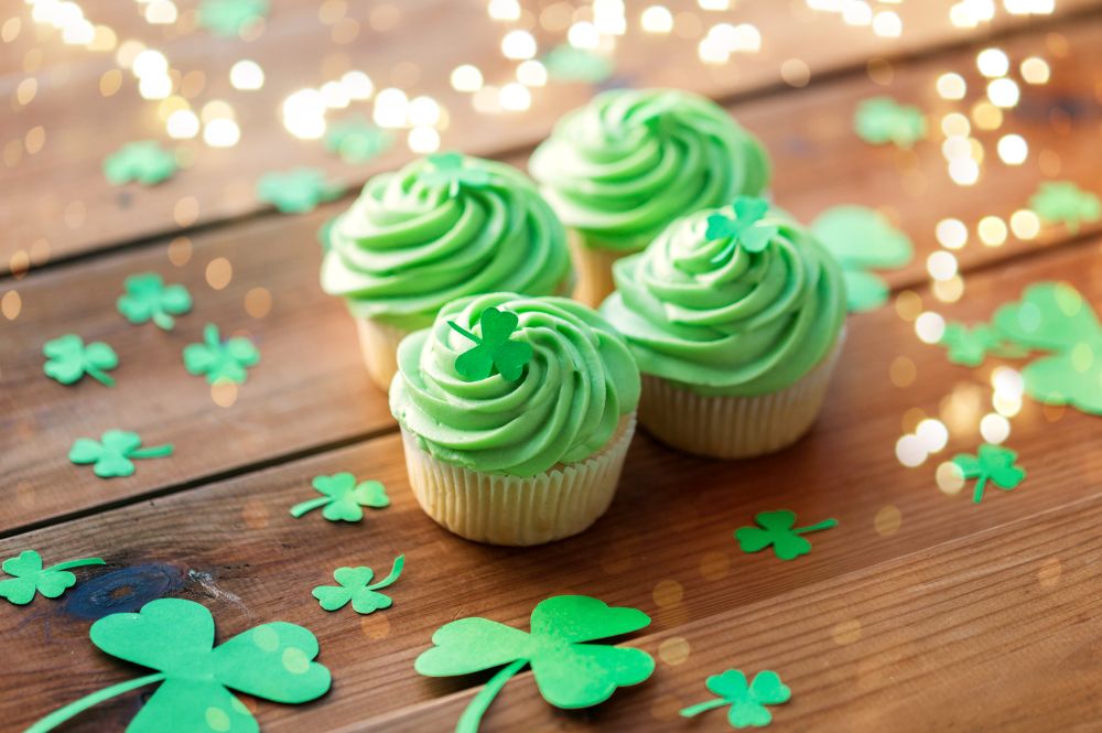 st patricks day, food and holidays concept - green cupcakes and shamrock on wooden table over festive lights. green cupcakes and shamrock on wooden table
