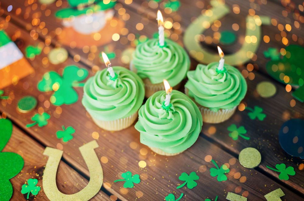 st patricks day, holidays and celebration concept - green cupcakes with candles and other party props on wooden table over festive lights. green cupcakes and st patricks day party props