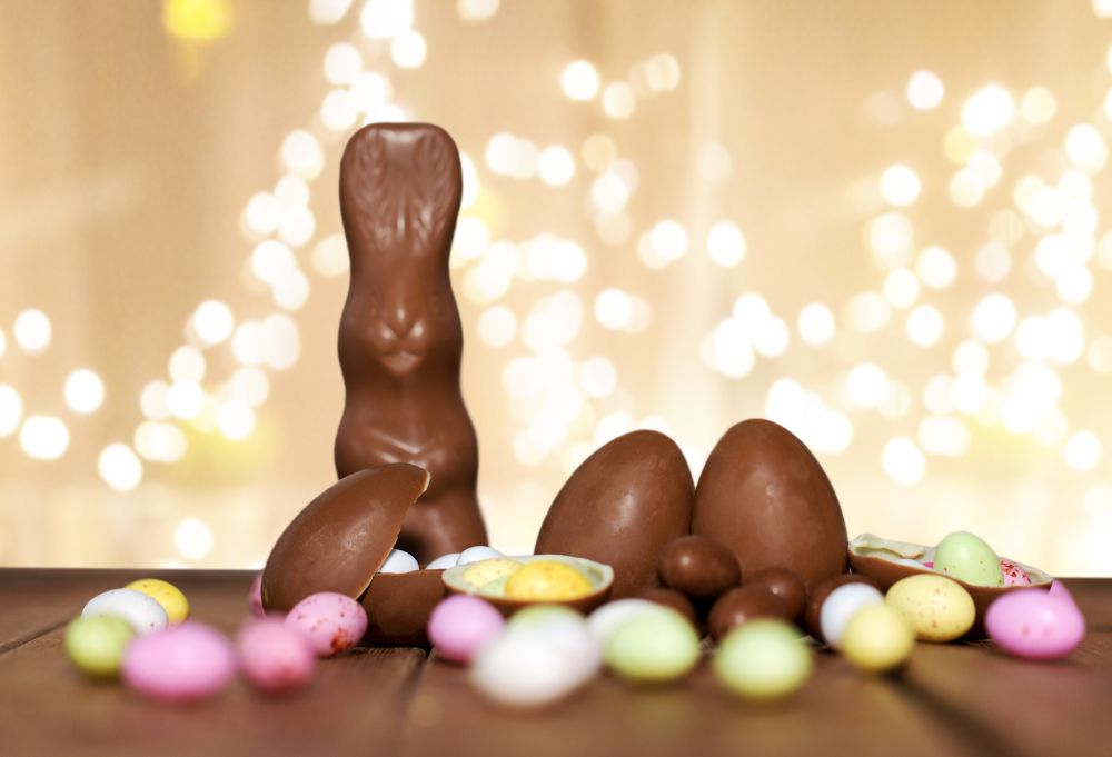 easter, sweets and confectionery concept - chocolate eggs, bunny and candy drops on wooden table over festive lights on beige background. chocolate eggs, easter bunny over festive lights