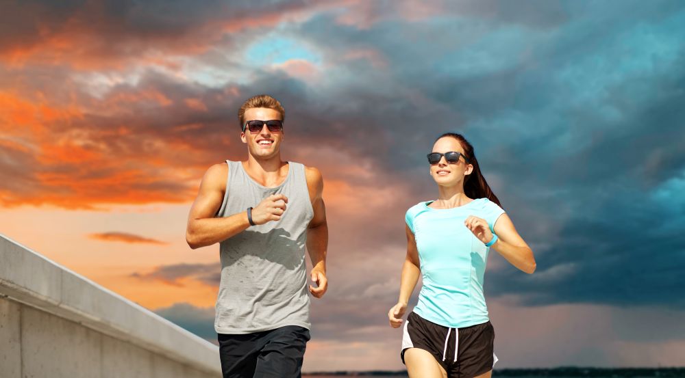 fitness, sport and healthy lifestyle concept - happy couple in sports clothes and sunglasses running over sunset sky background. couple in sports clothes running outdoors