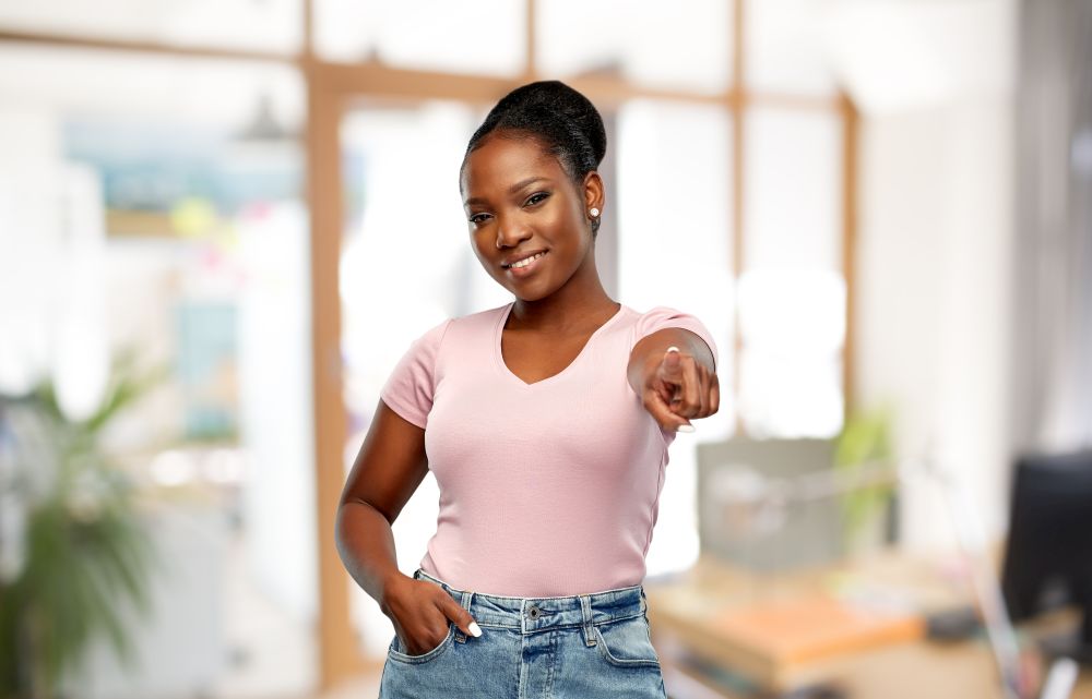 people, choice and gesture concept - happy african american young woman pointing finger to you over office background. african american woman pointing finger to you