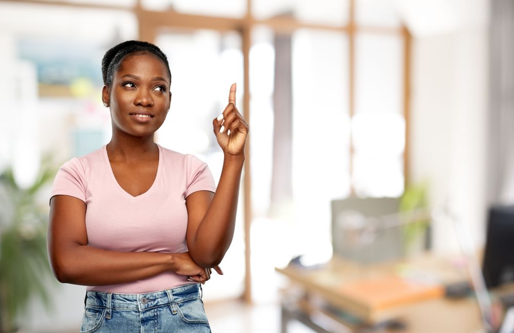 people, idea and attention concept - happy african american young woman pointing finger up over office background. happy african american woman pointing finger up