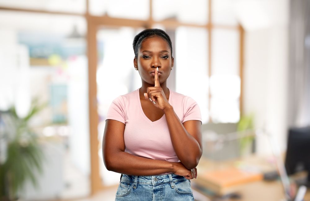 people, privacy and corporate concept - african american young woman with finger on lips over office background. african woman with finger on lips at office