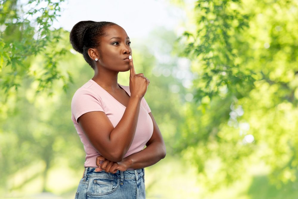 people, silence and secret concept - african american young woman with finger on lips over green natural background. african american woman with finger on lips