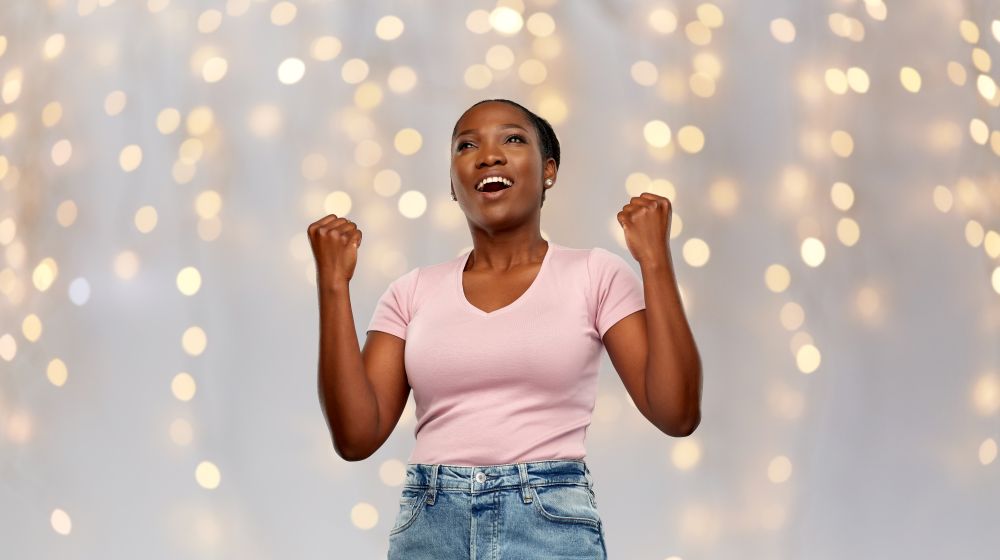 success, holidays and people concept - happy smiling young african american woman celebrating victory over festive lights background. happy african american woman celebrating success