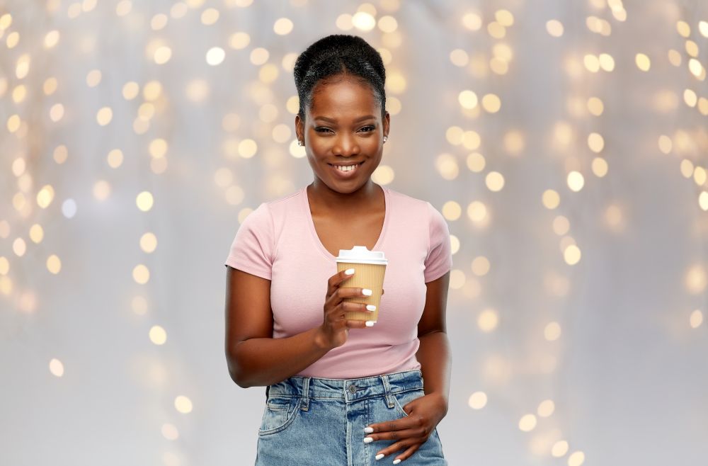 people, holidays and drinks concept - happy african american young woman drinking takeaway coffee from paper cup over festive lights background. happy african american woman drinking coffee