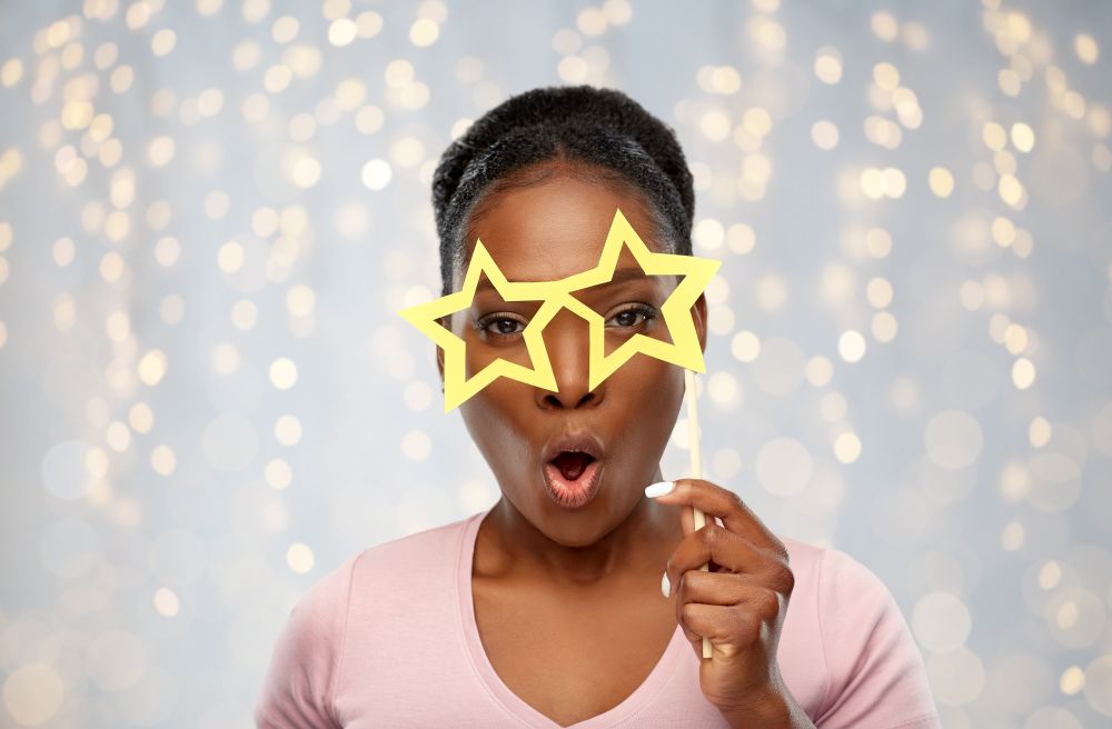 party props, photo booth and people concept - surprised african american young woman with star shaped glasses over festive lights background. african american woman with star shaped glasses