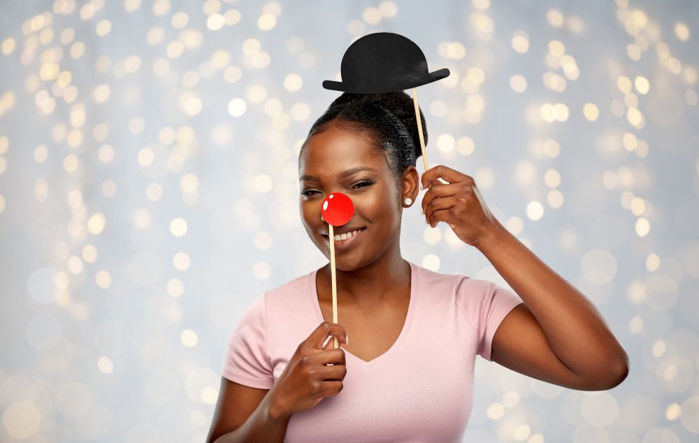red nose day, party props and photo booth concept concept - happy african american young woman with clown nose over festive lights background. happy african american woman with red clown nose