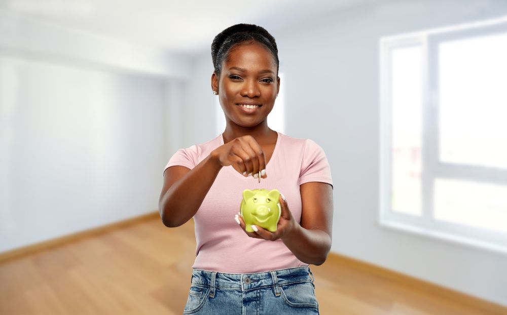 mortgage, saving and finance concept - happy smiling african american young woman with piggy bank over empty home room background. african american woman with piggy bank at new home