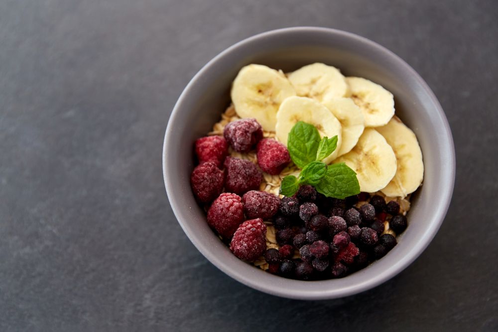 food and breakfast concept - oatmeal cereals in bowl with wild berries, banana and peppermint on slate stone table. cereal breakfast with berries, banana and mint