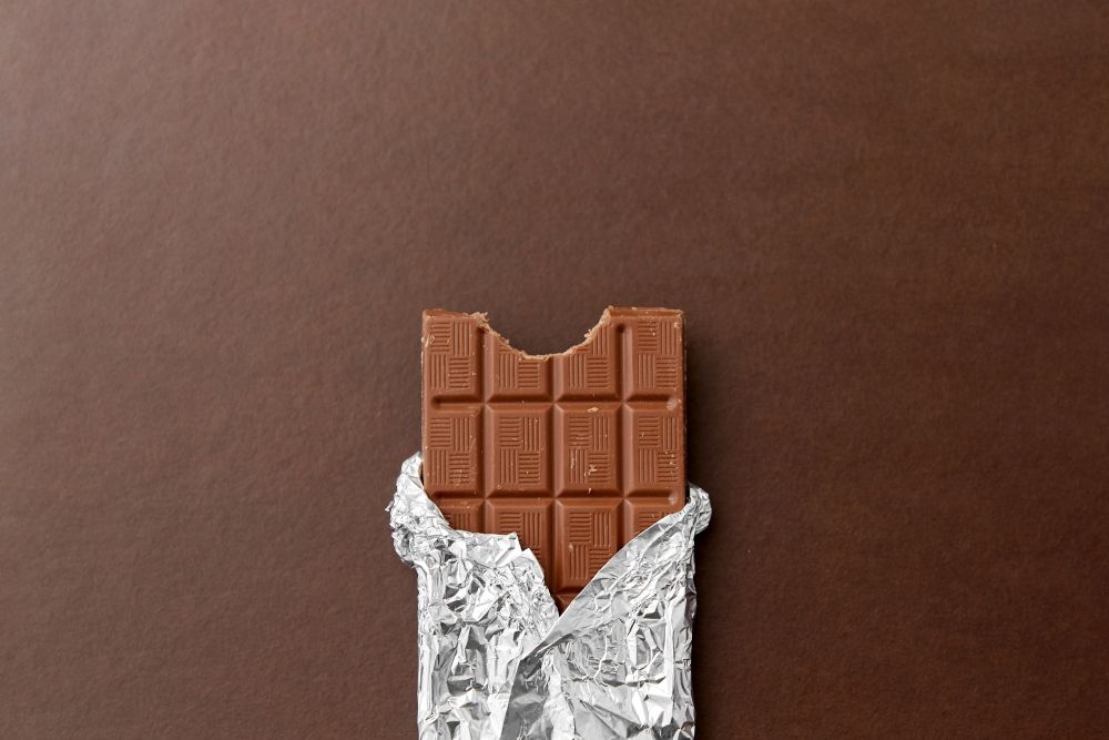 sweets, confectionery and food concept - milk chocolate bar in foil wrapper on brown background. chocolate bar in foil wrapper on brown background