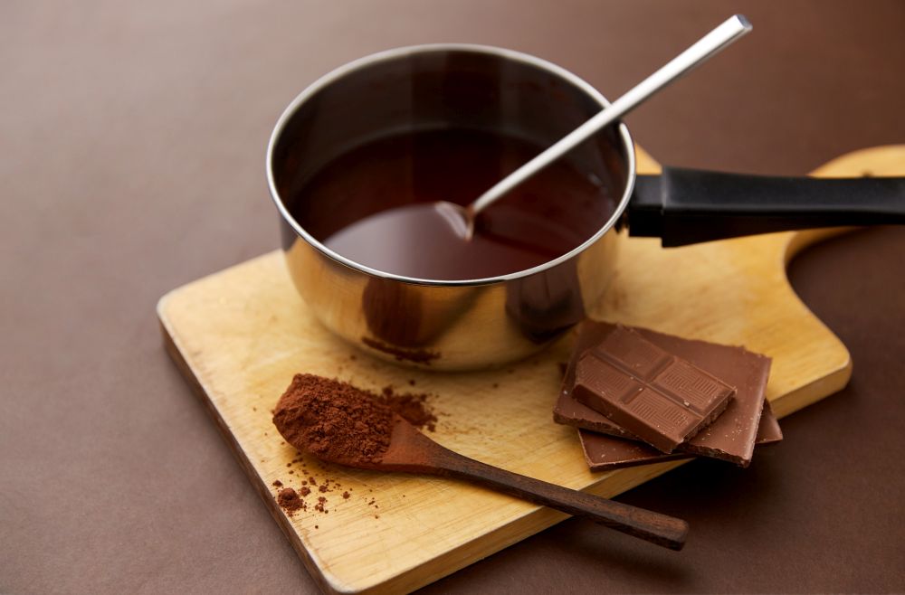 sweets, confectionery and culinary concept - pot with melted hot chocolate, cocoa powder in spoon and wooden board on brown background. pot with melted hot chocolate and cocoa powder